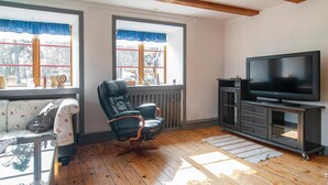 A living room with a black leather chair, a sofa with floral upholstery, a television on a dark wooden stand, and large windows with blue curtains.