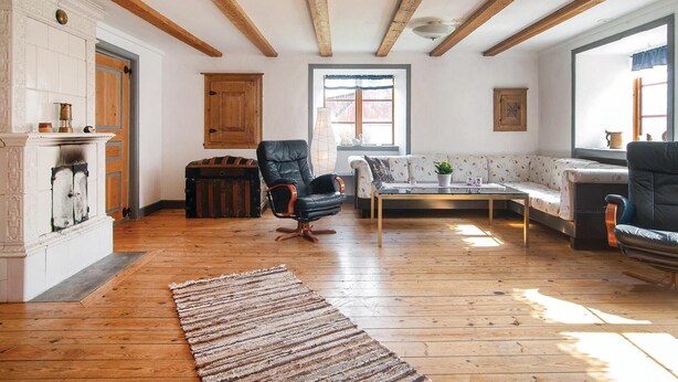 A spacious living room with wooden floors, a fireplace, a black leather armchair, a floral sofa, and a large window providing natural light.