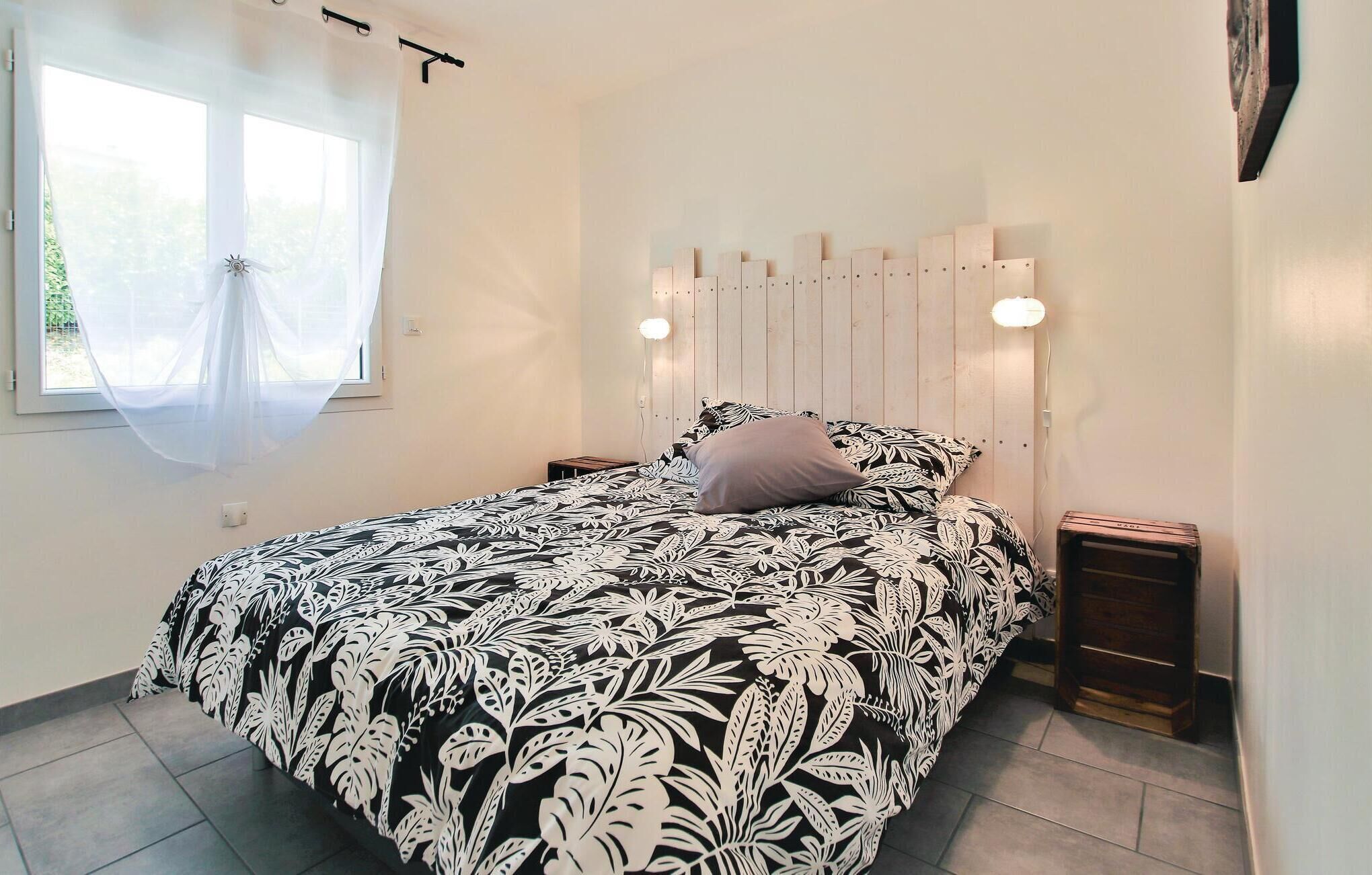 Un dormitorio con cama, mesita de noche, ventana con cortinas traslúcidas y lámpara de pared.