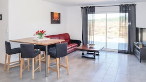 Ein modernes Wohnzimmer mit einem roten Ledersofa, einem Holzesstisch mit vier Stühlen und einem Couchtisch. An der Wand hängt ein Gemälde und ein Fenster mit Vorhängen.