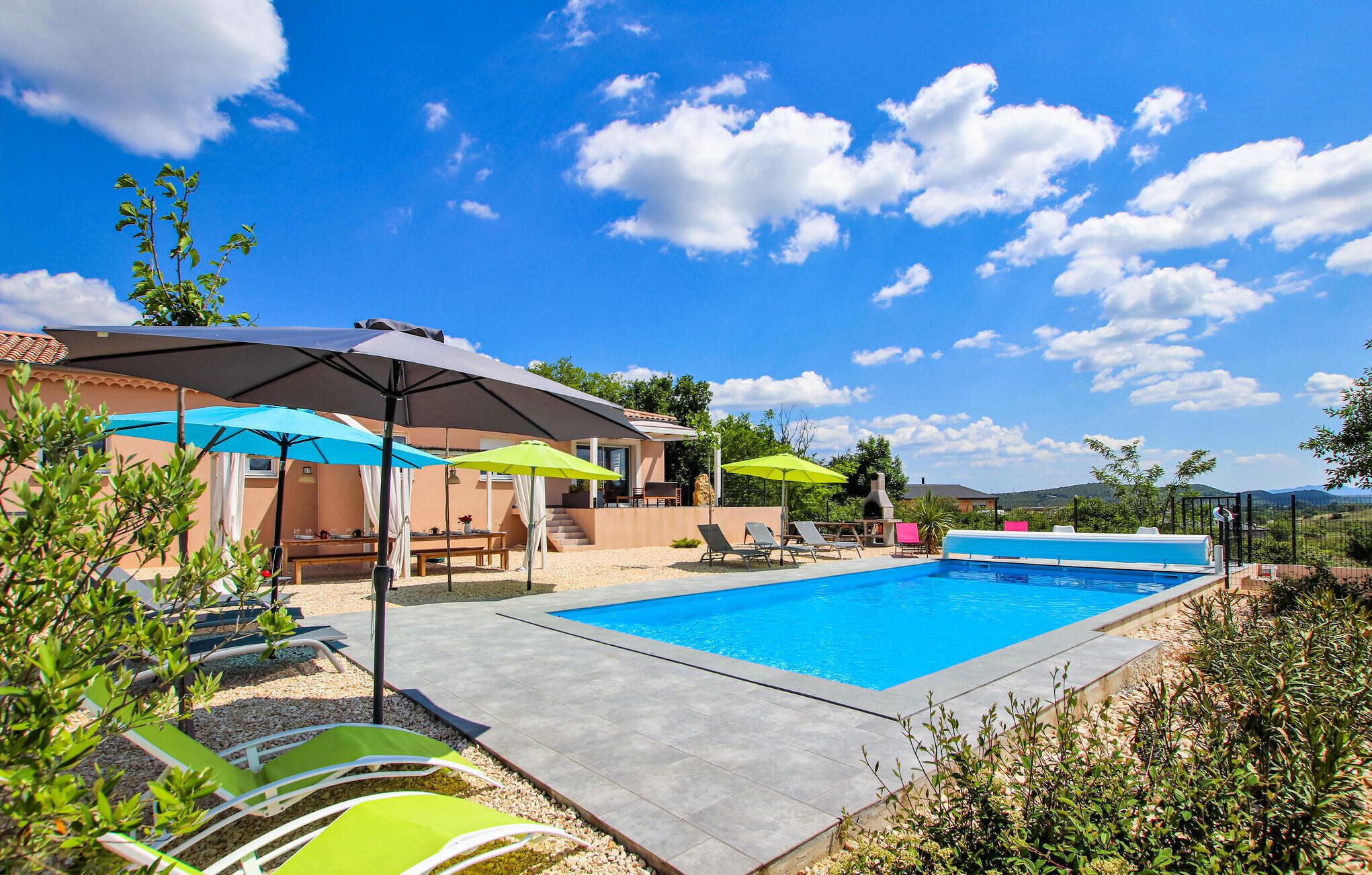A swimming pool with lounge chairs and umbrellas, surrounded by a landscaped garden and a building with a tiled roof.