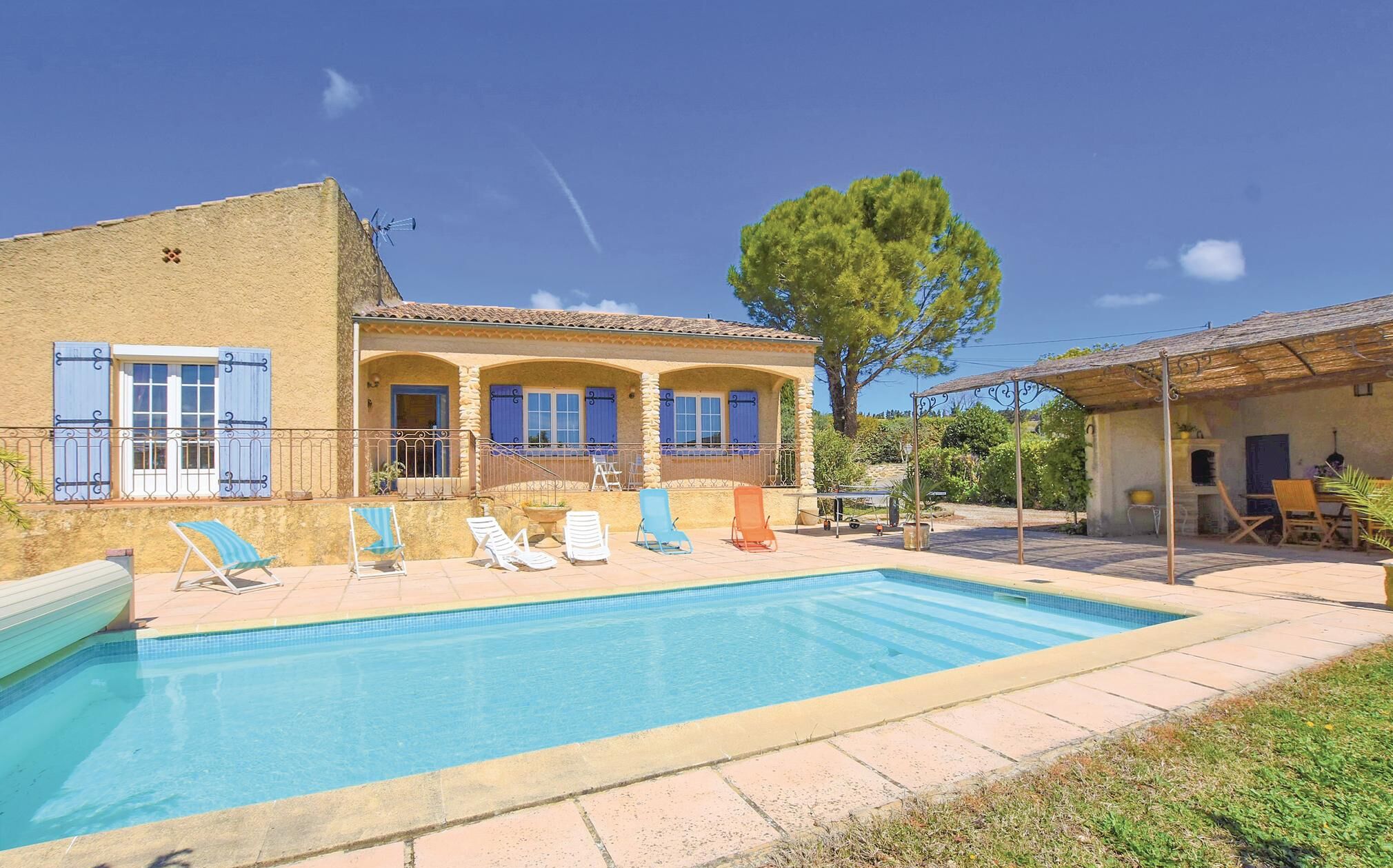 A poolside area with a pool, lounge chairs, and a building with a tiled roof and blue shutters.