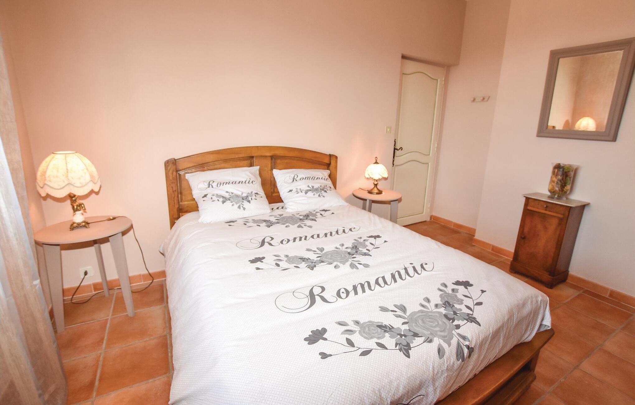 Une chambre à coucher avec un lit recouvert d’une couvre-lit à motifs floraux, orné du mot « Romantique », et agrémentée de coussins assortis. On y trouve une table de chevet avec une lampe, ainsi qu’une petite table également dotée d’une lampe. Un meuble en bois est adossé au mur.