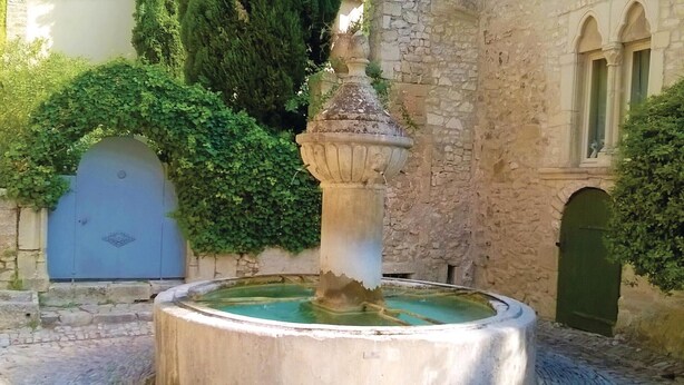 A stone fountain with water in a courtyard, surrounded by stone walls and greenery.