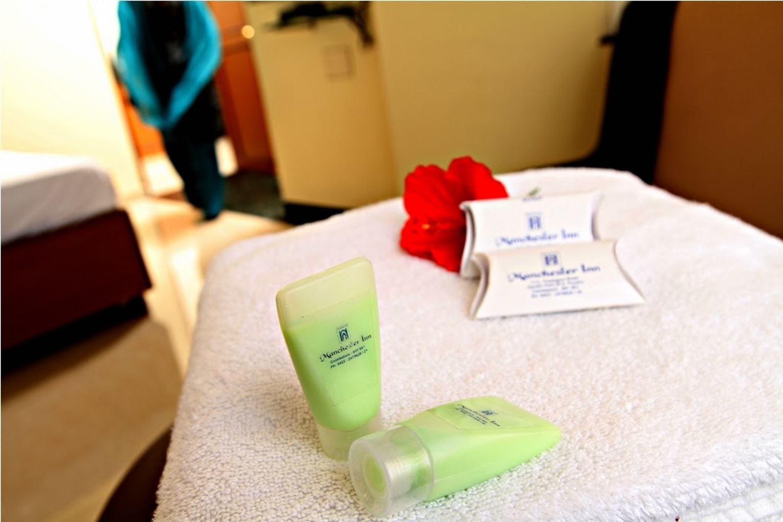 Een hotelkamer met een bed, een witte handdoek en een rode roos. Op het bed staan twee groene flesjes, waarvan één een fles met lotion is. Een witte kaart met de naam van het hotel en contactgegevens is ook zichtbaar.