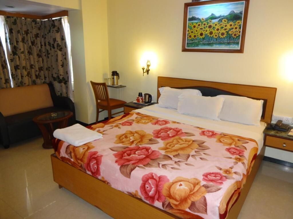 Quarto de hotel com uma cama grande, cabeceira de madeira, colcha florida, uma cadeira, uma mesinha e um quadro de girassóis, além de uma janela com cortinas.