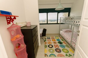 A small, well-lit room with a bunk bed, a desk with drawers, a chair, a hanging pink storage organizer, and a window with a view.