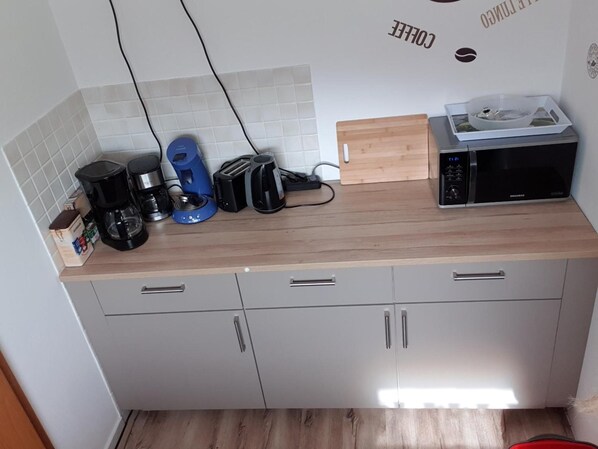 Una cocina con microondas, cafetera, tostadora y una tabla de cortar de madera sobre la encimera.