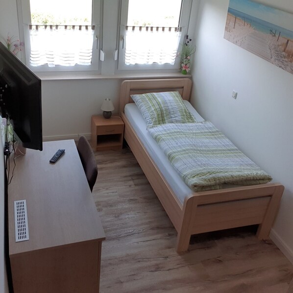 A bedroom with a bed, a desk, a chair, a small bedside table with a lamp, and a window with blinds. - Gästehaus Strudthoff (Ganderkesee)