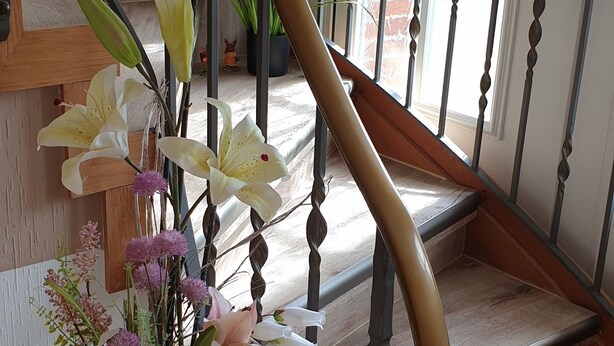 Escada de madeira com corrimão de metal, um vaso com flores e um muro de tijolos ao fundo.