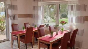 Uma área de refeições com uma mesa posta para seis pessoas, rodeada de cadeiras, e vista para um jardim através de grandes janelas.