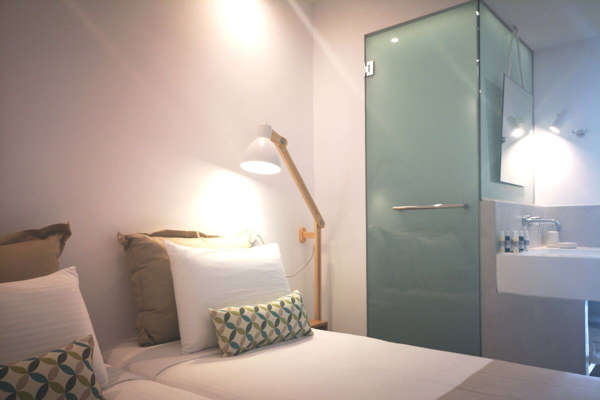 Une chambre d’hôtel avec un lit, des oreillers, une lampe de chevet et une porte en verre donnant sur une salle de bain équipée d’un lavabo et d’un miroir.
