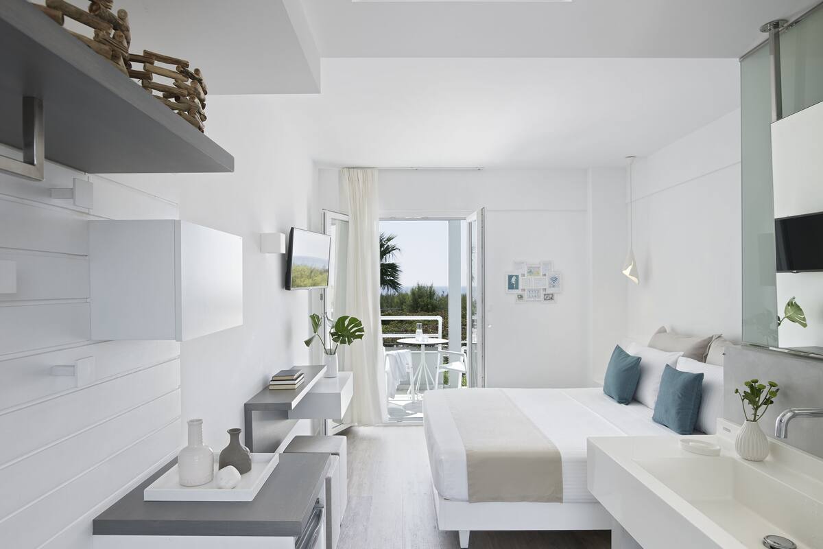 Une chambre d’hôtel moderne au design minimaliste, comprenant un lit, un grand miroir, un bureau intégré et une vue sur l’extérieur.