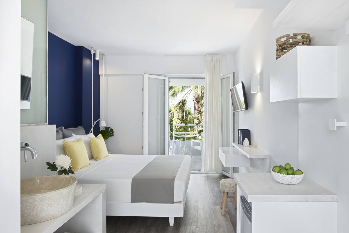 Une chambre d’hôtel moderne, équipée d’un lit, d’une salle de bain avec un lavabo et un miroir, d’un petit bureau avec un bol de fruits, et offrant une vue sur l’extérieur.