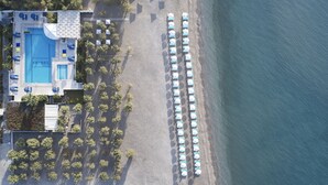Vista aérea de un hotel frente a la playa con piscina, sombrillas y una fila de sillas.