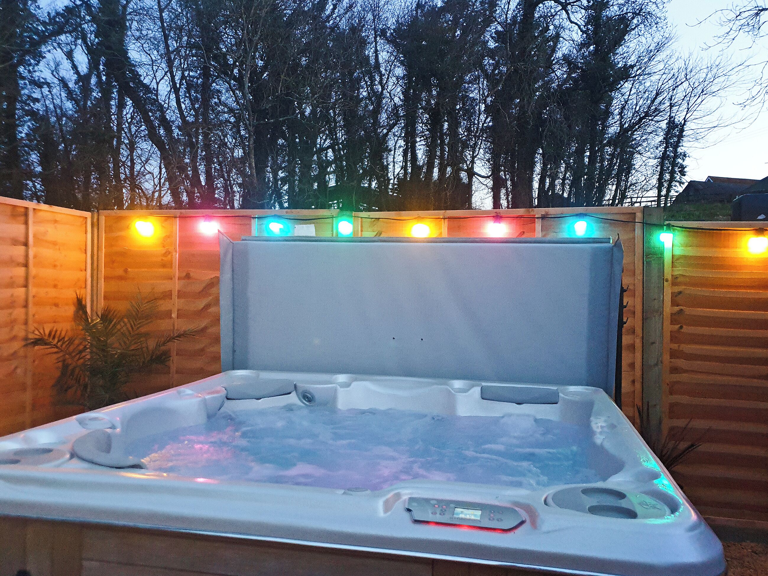 Un bain à remous doté d’un panneau de commande, entouré d’une clôture en bois et illuminé de guirlandes lumineuses.