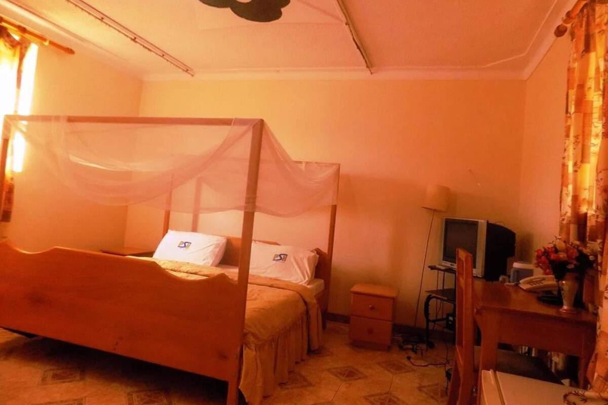 A room with a four-poster bed, a desk with a chair, a television, and a small bedside table.