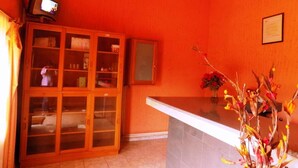 A room with orange walls, a wooden cabinet with glass doors, a small television on top, and a vase with flowers on a counter. - Palm Springs Hotel (Masaka)