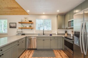 Une cuisine moderne avec des meubles gris, des appareils en acier inoxydable, un lave-vaisselle et une fenêtre équipée de stores.