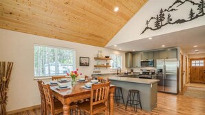 A dining area with a wooden table and chairs, a kitchen with modern appliances, and a large window with a view of trees.