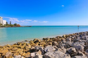 Une côte rocheuse bordée d’une eau turquoise cristalline, avec une jetée et des bâtiments en arrière-plan.