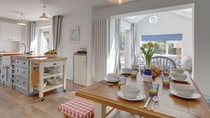 A bright kitchen with a wooden table set for breakfast, a white cabinet, and a view through a large window. - Egret, South Creake, Norfolk (South Creake)