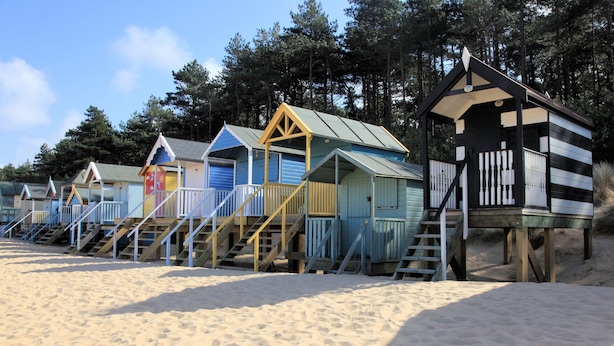 A row of beach huts with wooden stairs leading up to them, set on sandy ground with trees in the background. - Egret, South Creake, Norfolk (South Creake)