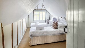 Quarto com teto inclinado, janela com persianas, duas camas com roupa de cama branca e um tapete listrado.