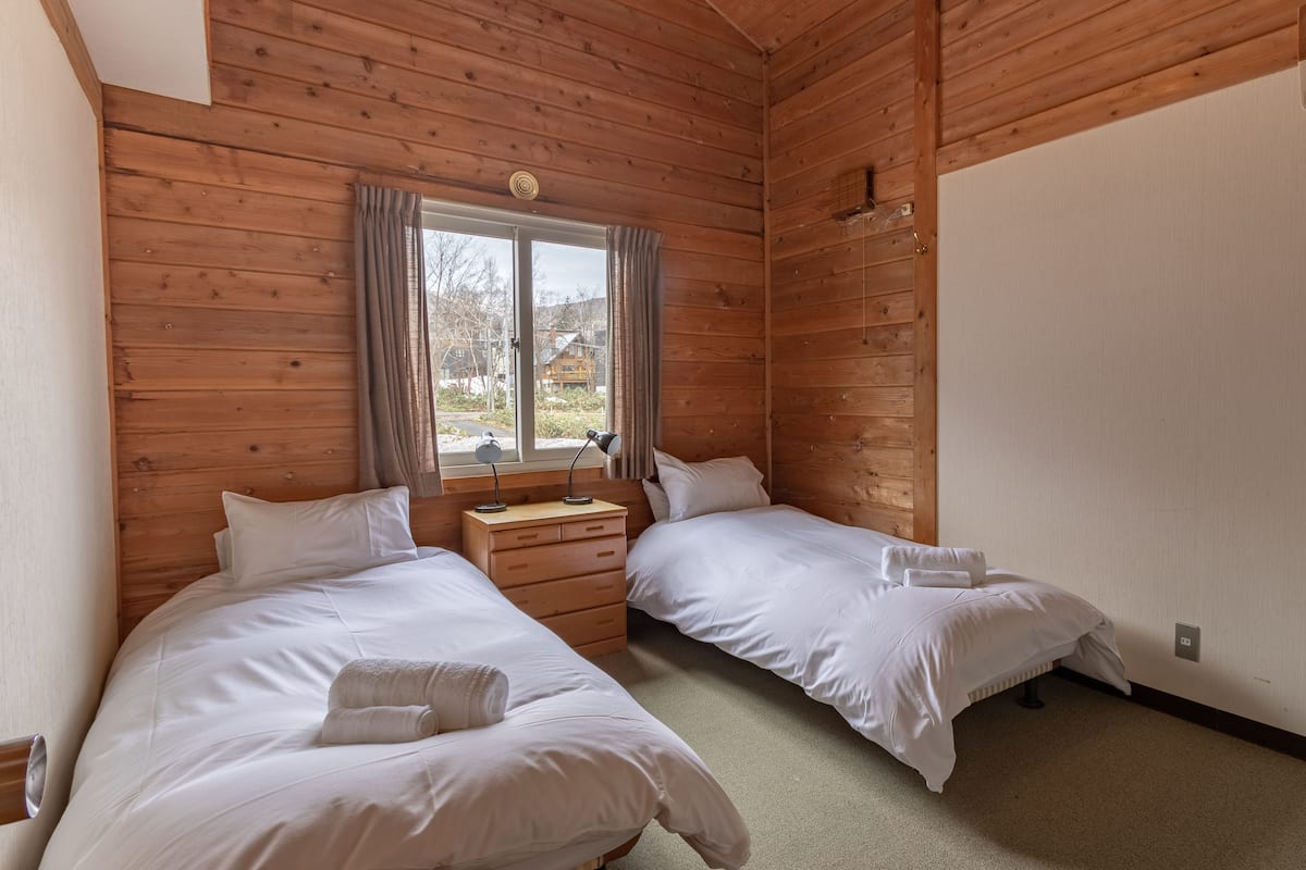 Una habitación con paneles de madera, dos camas, una mesita de noche y una ventana con cortinas.
