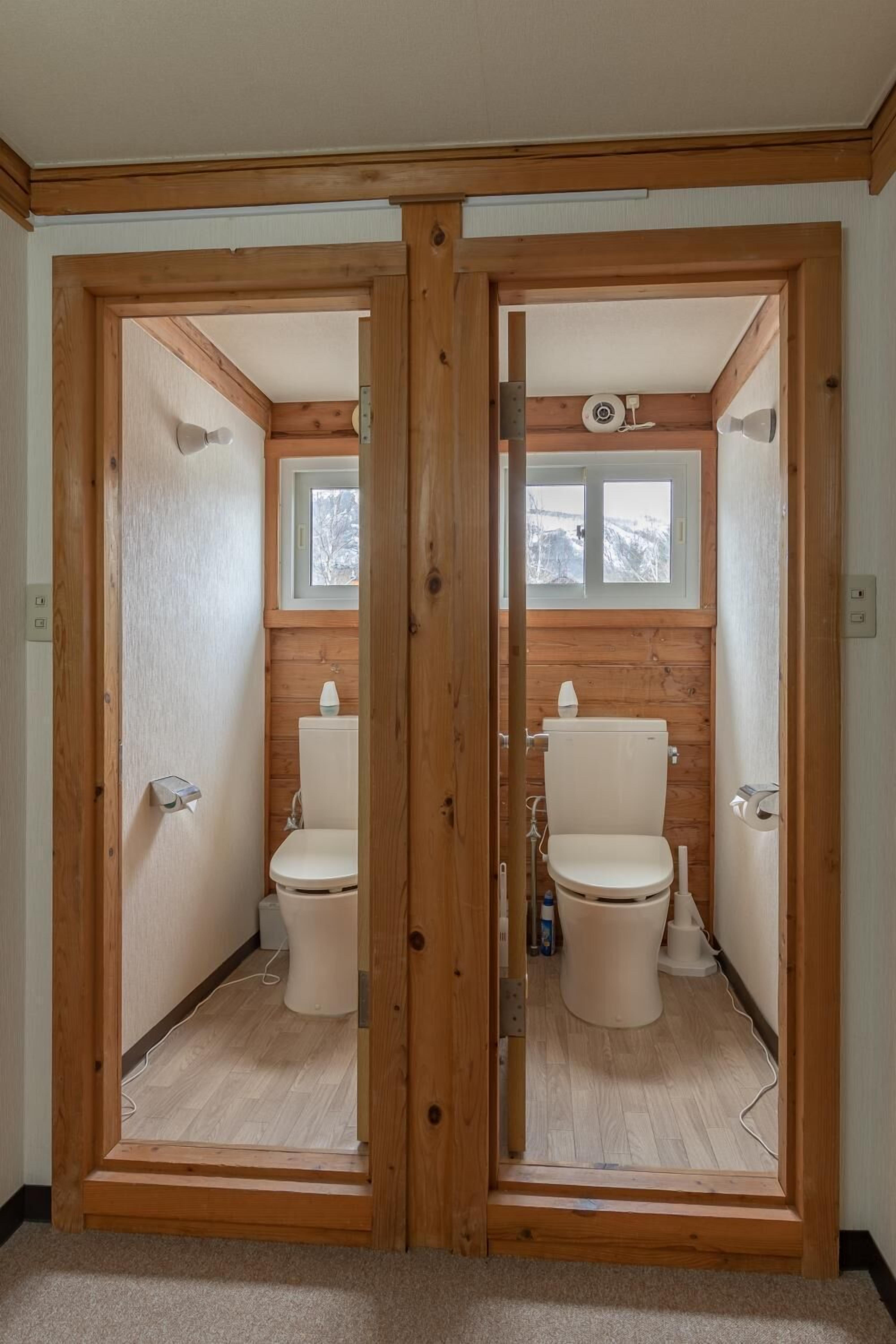 A wooden-framed bathroom with two toilets, a window above the sink, and a light fixture on the wall.