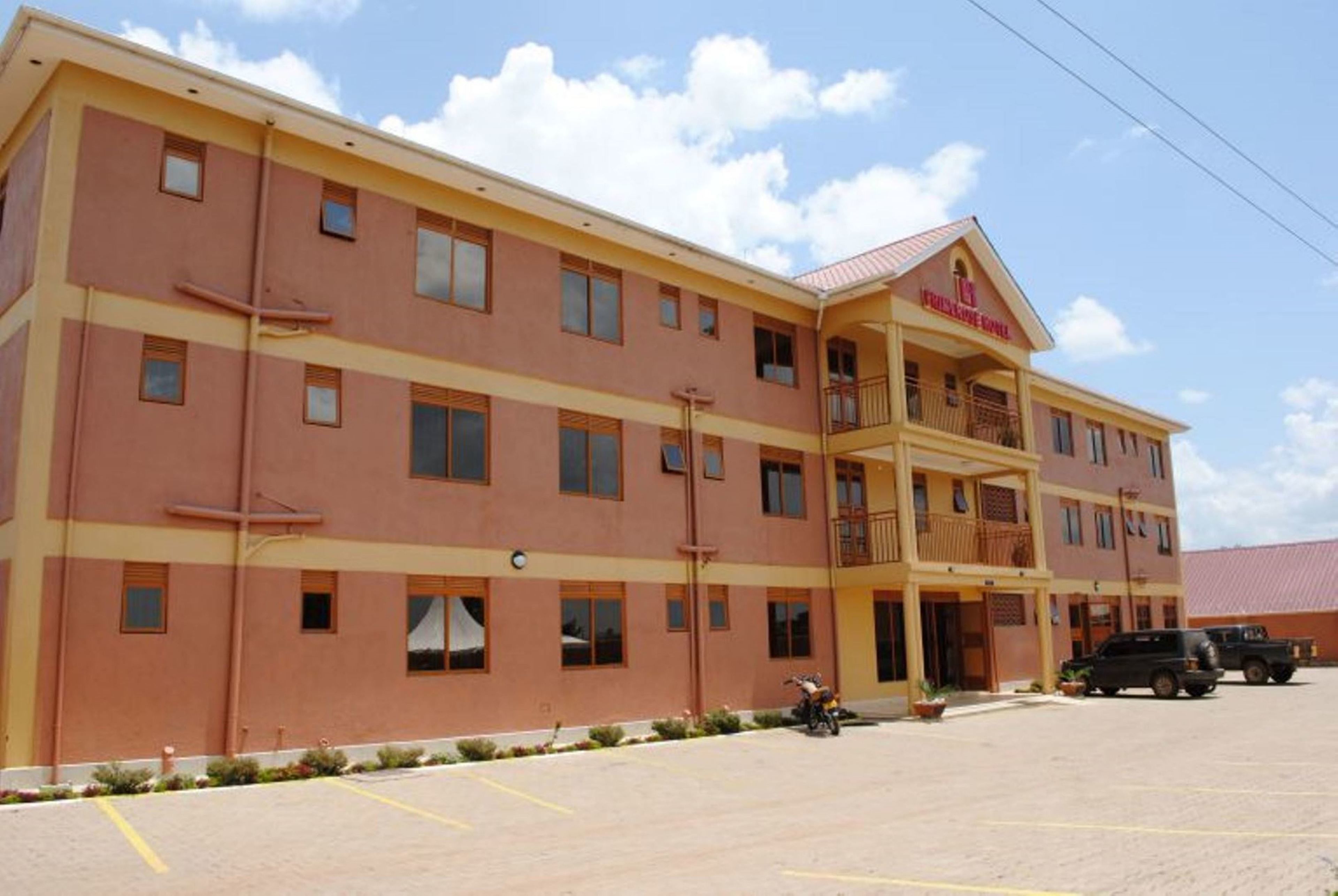 A multi-story hotel with a yellow facade, a balcony on the second floor, and a parking lot with two vehicles.