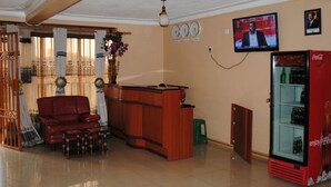 Una sala de estar con un mostrador de recepción de madera, un sofá rojo, un taburete verde, un televisor montado en la pared y un refrigerador rojo Coca-Cola.