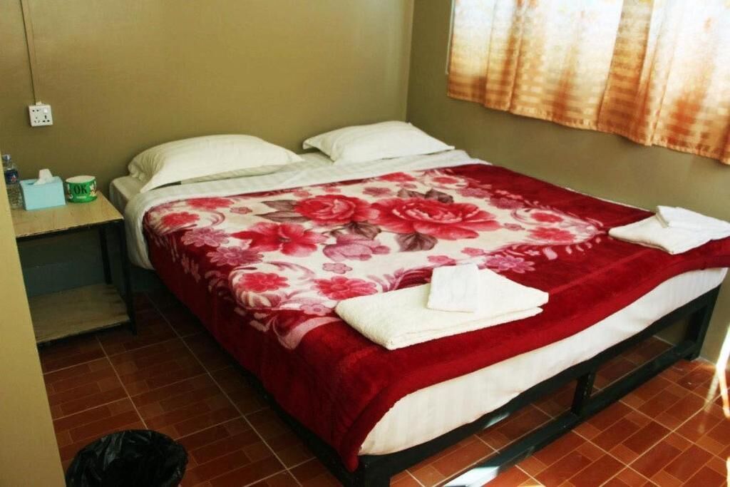 Uma cama de solteiro com colcha florida, uma mesinha de cabeceira com caixa de lenços e um recipiente verde, e um saco de lixo preto no chão.
