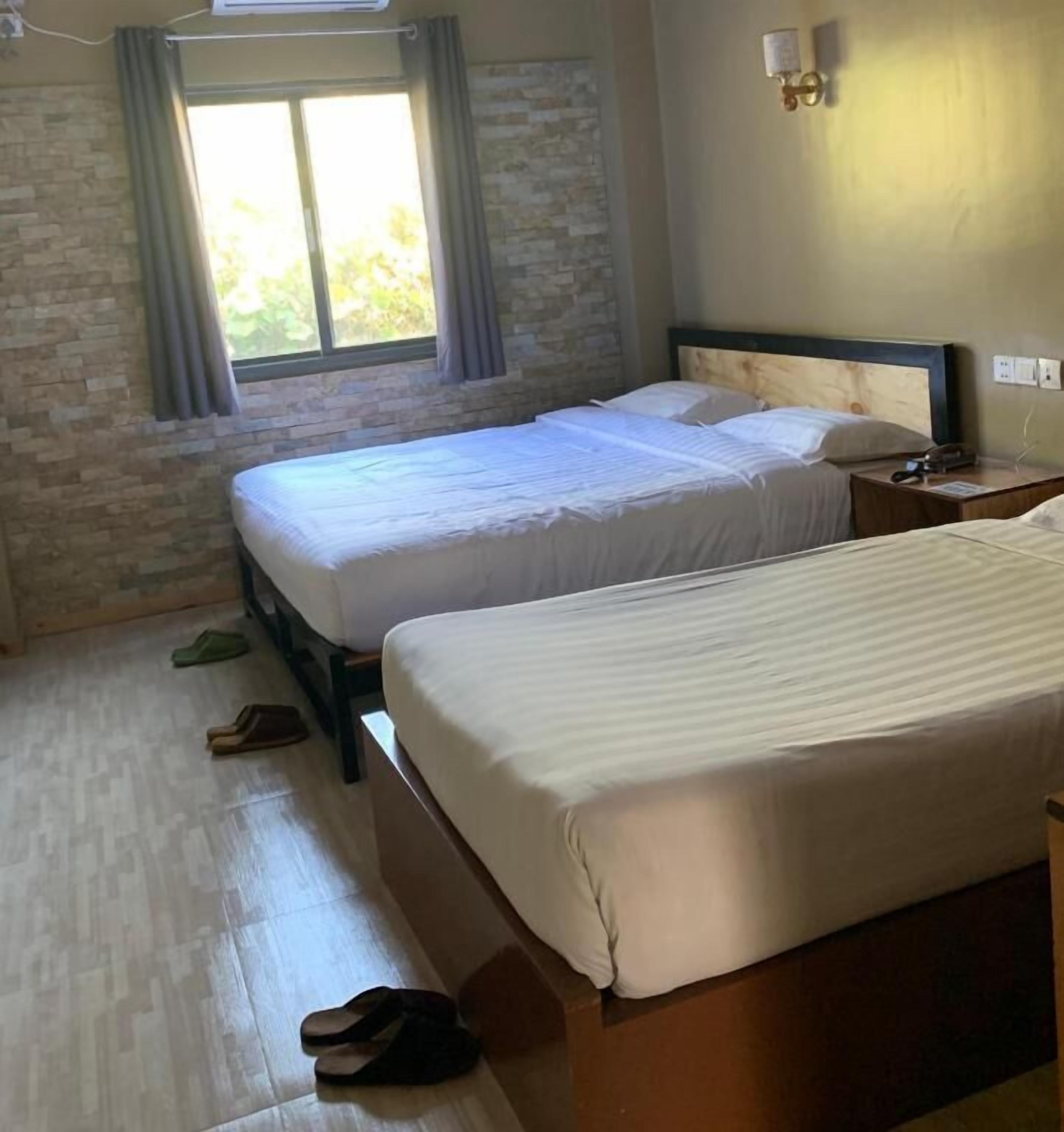 A hotel room with two beds, a stone wall, a window with curtains, a nightstand with a lamp, and slippers on the floor.