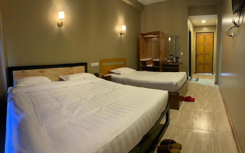 A hotel room with two beds, a wooden wardrobe, a small table with a lamp, and a door leading to another room.