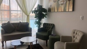 Olohuoneessa on sohva, kaksi nojatuolia, sohvapöytä ja suuri ruukussa oleva kasvi.
