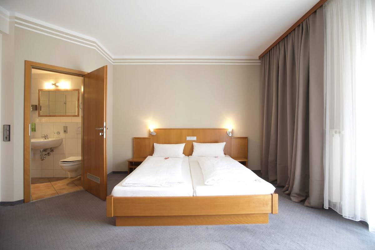 A hotel room with a double bed, a wooden headboard, a bathroom with a sink and mirror, and a large window with curtains.