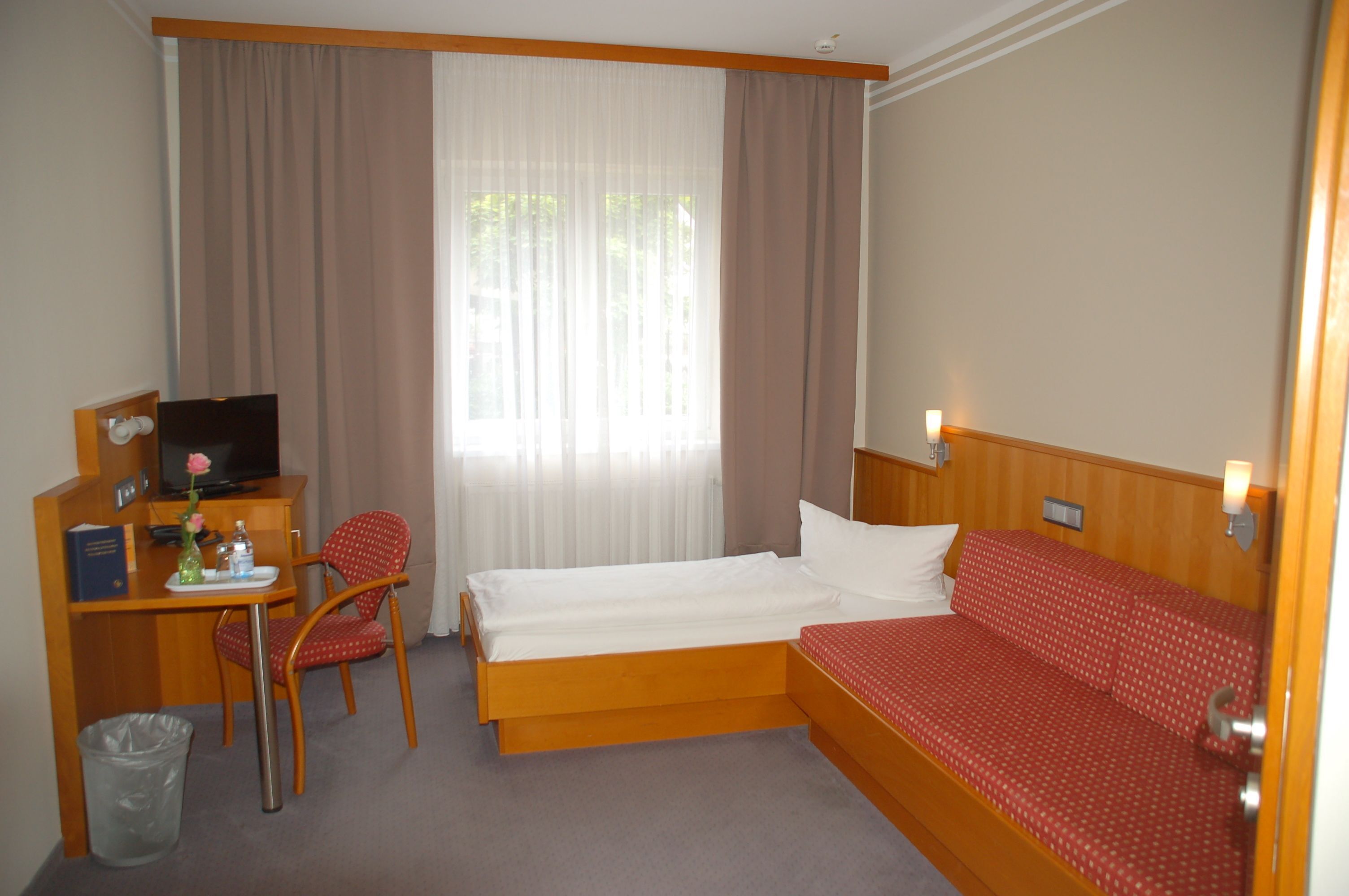 A hotel room with a bed, a desk with a TV, a chair, and a window with curtains.