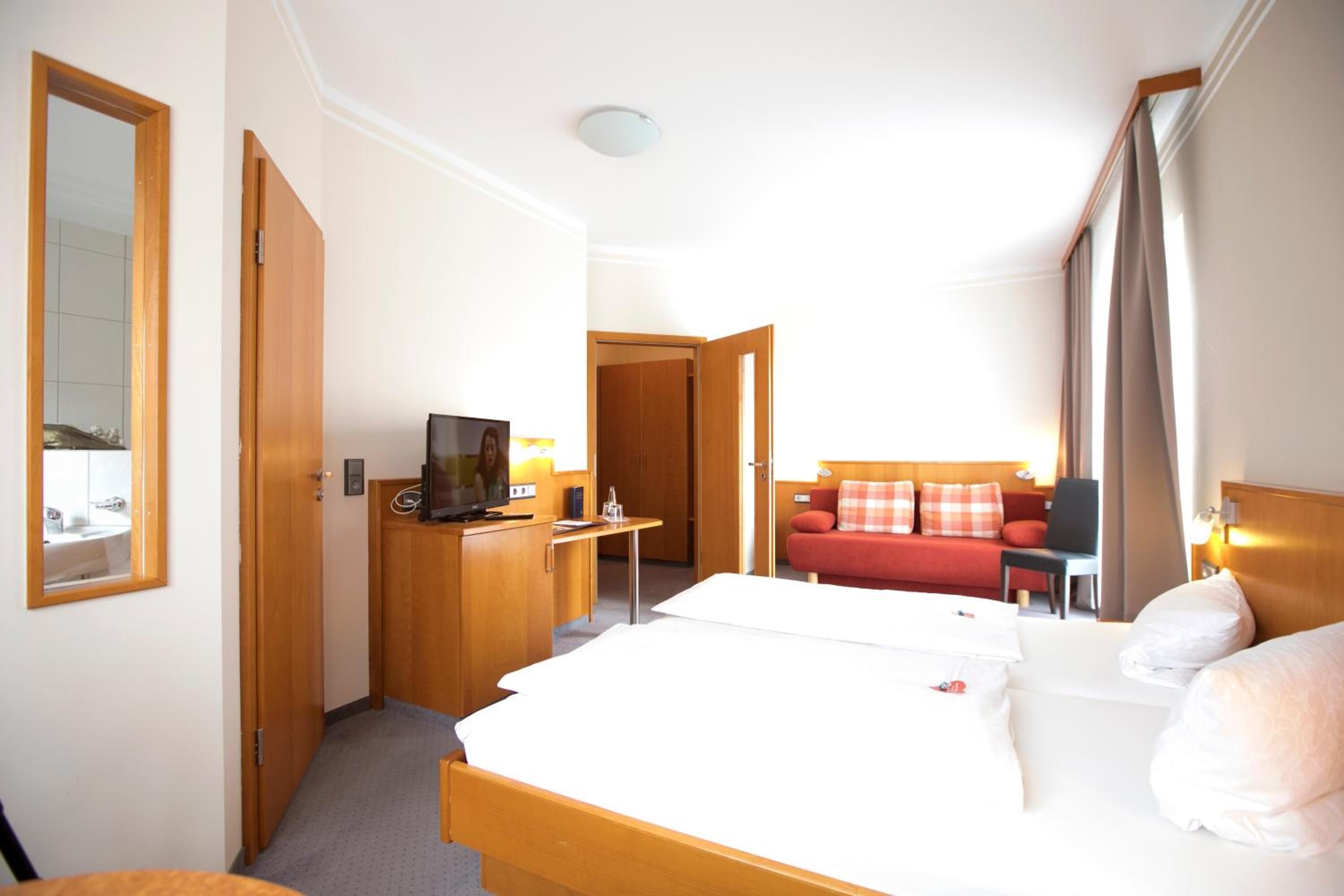 Quarto de hotel com cama, escrivaninha com televisão, sofá vermelho e espelho.