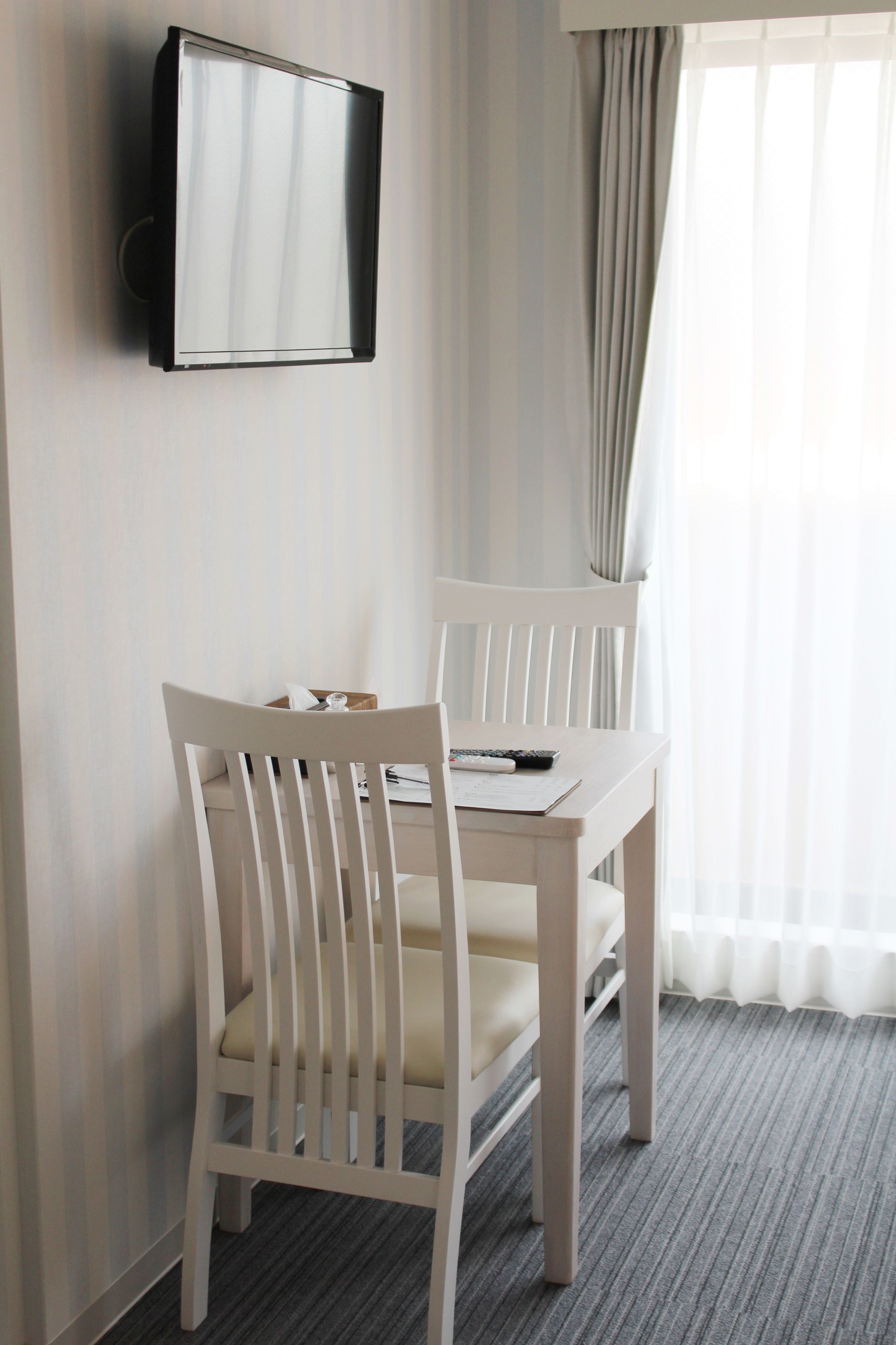 Hotelkamer met een aan de muur bevestigde televisie, twee witte stoelen, een klein tafeltje en een raam met gordijnen.