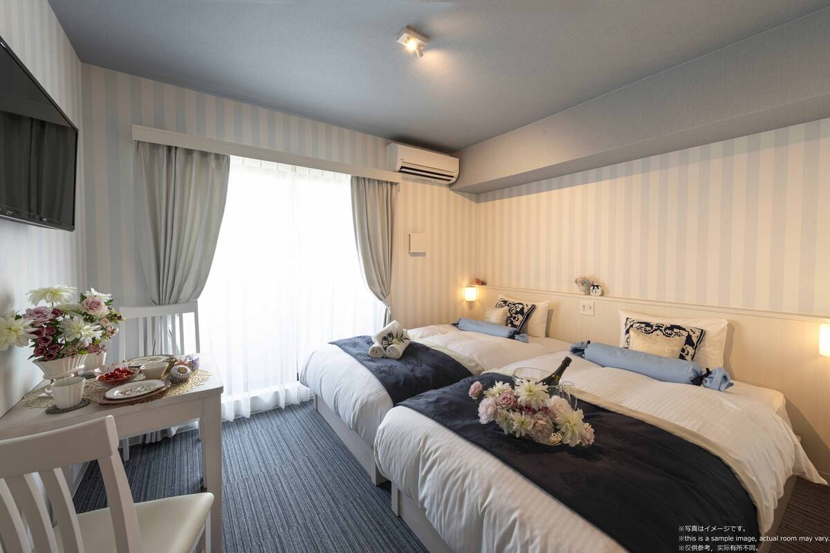 A hotel room with two beds, a desk with a chair, a flat-screen TV, and a window with curtains.