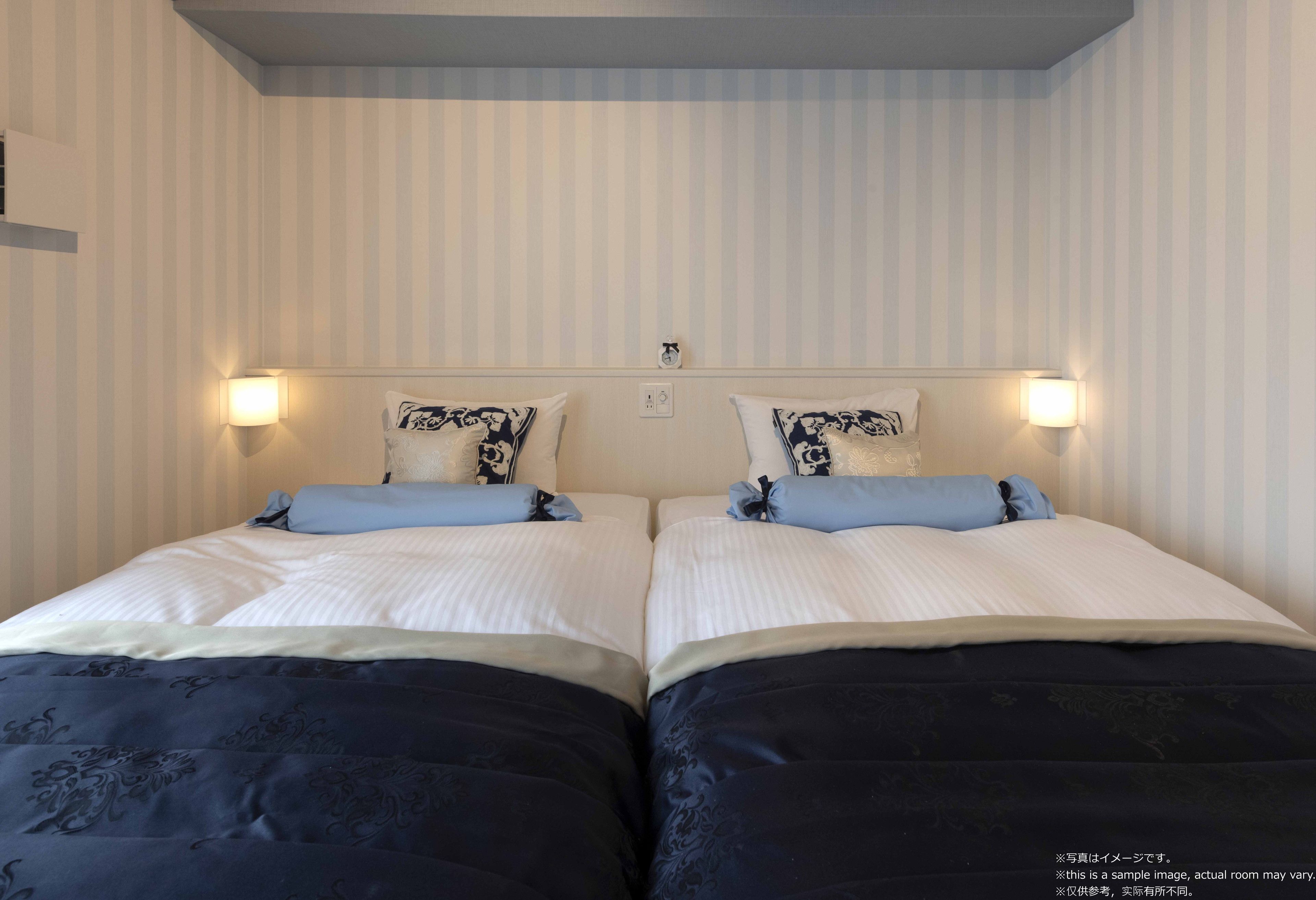 A double bed with a white and blue bedspread, two matching pillows, and two wall-mounted night lights.