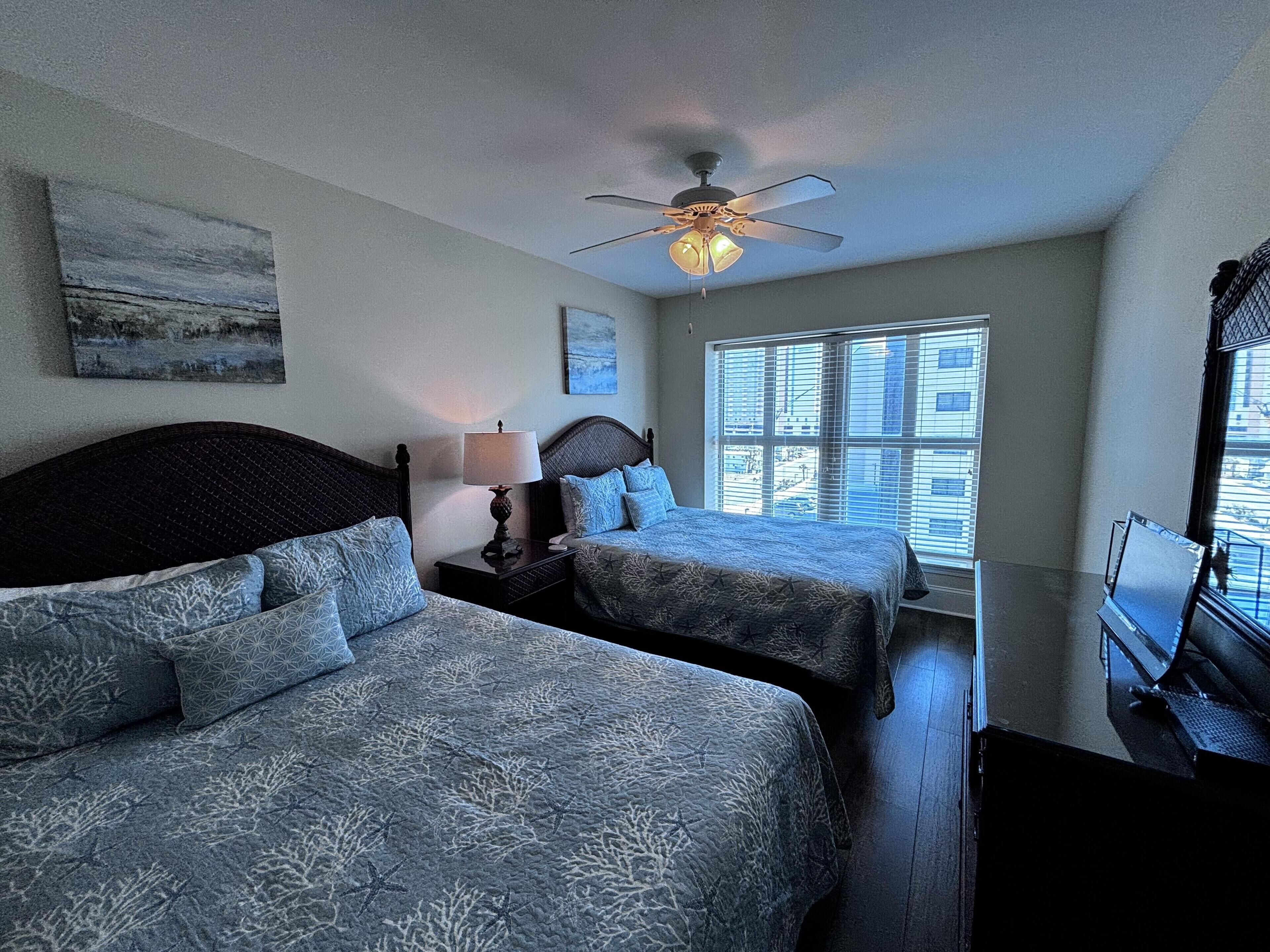A bedroom with two beds, a ceiling fan, a nightstand with a lamp, and a window with blinds.