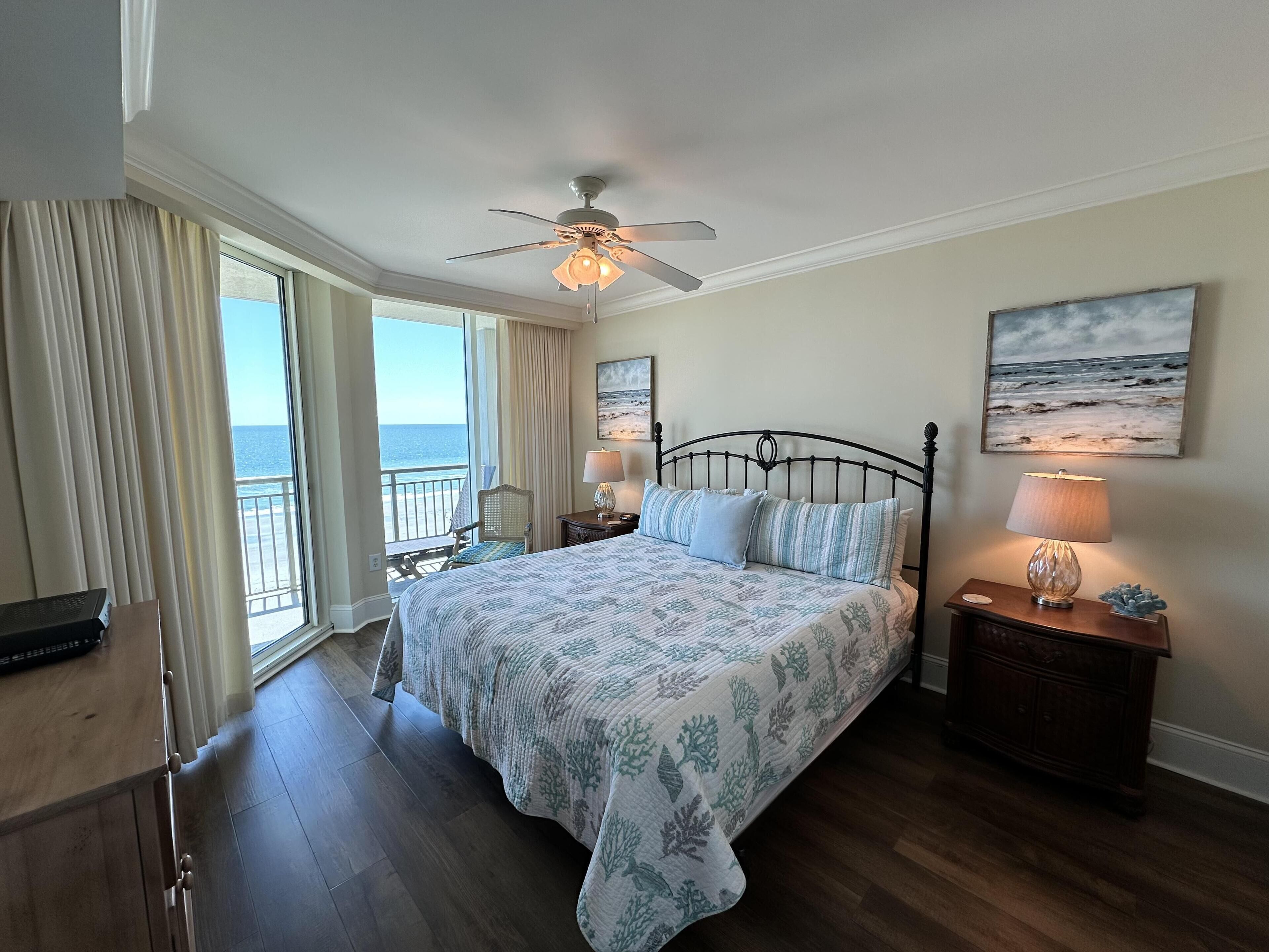 A bedroom with a bed, a ceiling fan, a nightstand with a lamp, a chair, a view of the ocean, and a painting on the wall.