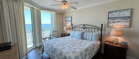 A bedroom with a bed, a ceiling fan, a nightstand with a lamp, a chair, a view of the ocean, and a painting on the wall.