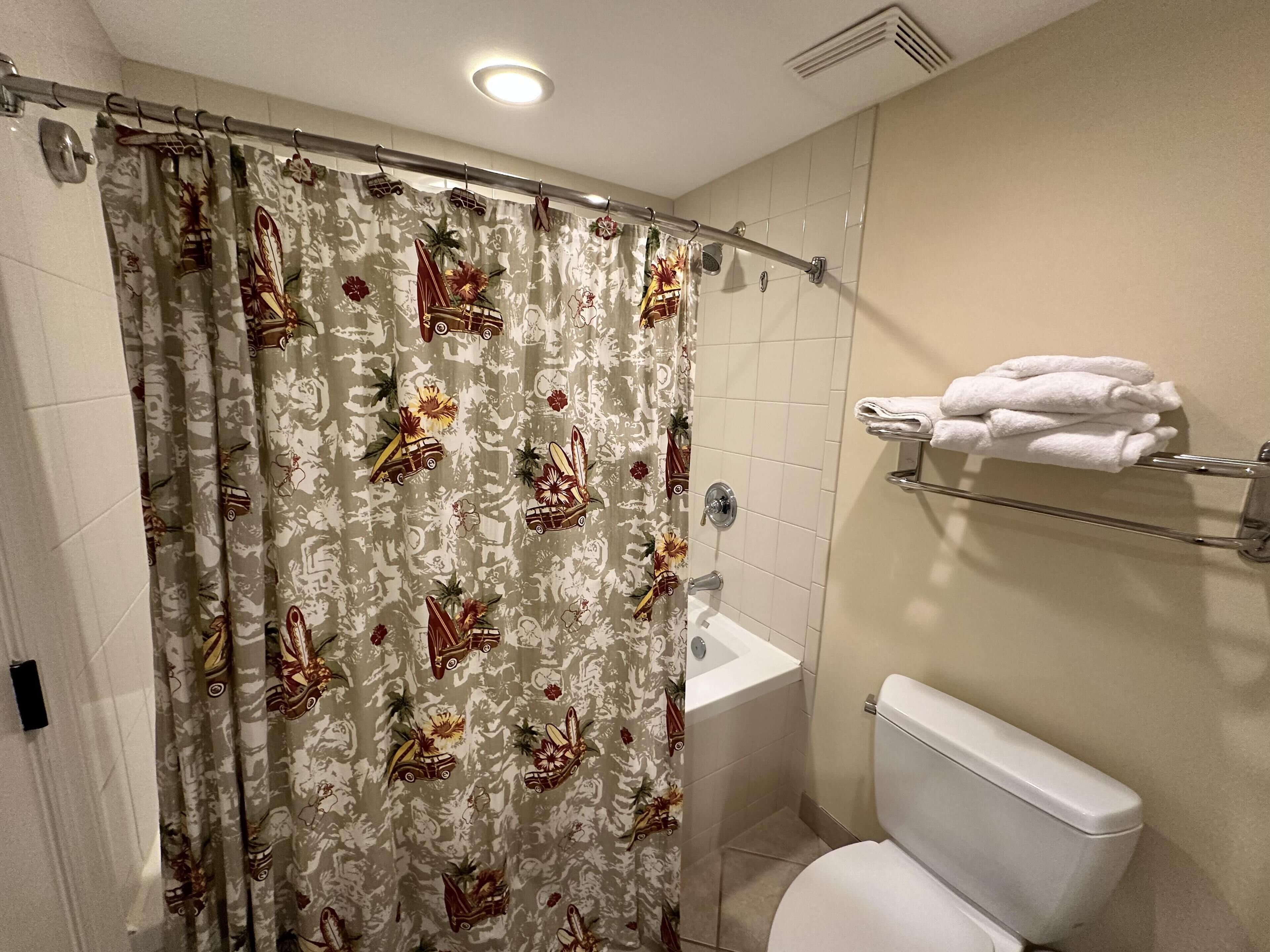 A bathroom with a shower curtain featuring a tropical pattern, a white bathtub, and a toilet with a towel rack above it.