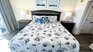 A bedroom with a bed featuring a nautical-themed bedspread, two framed pictures on the wall, and a nightstand with a lamp.