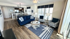 A living room with a blue sofa, patterned chairs, a coffee table, and a dining area with a kitchen in the background.