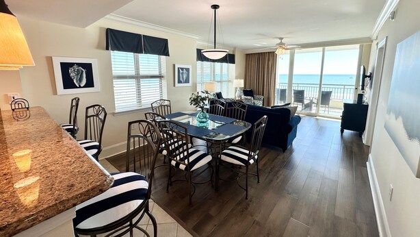 A modern kitchen with a granite countertop, dining area with striped chairs, a sofa, and a balcony with a view of the ocean.