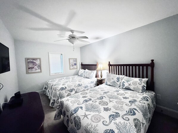 Une chambre avec deux lits, un ventilateur de plafond, une table de chevet avec une lampe et un téléviseur fixé au mur.